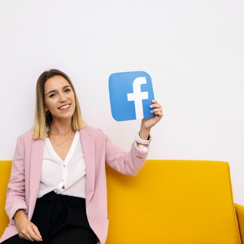 smiling-young-woman-sitting-yellow-sofa-holding-facebook-icon.jpg